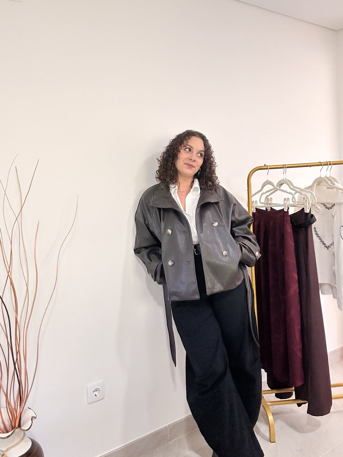 Silhouette en veste Noelle en simili cuir marron, associée à une chemise blanche et un pantalon noir, en intérieur.